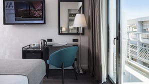Habitación de hotel con cama, escritorio, silla, televisión, espejo, lámpara y balcón con barandilla.