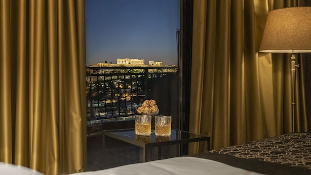 Habitación de hotel con vistas a arquitectura griega antigua, una mesita de noche con dos vasos y una lámpara.