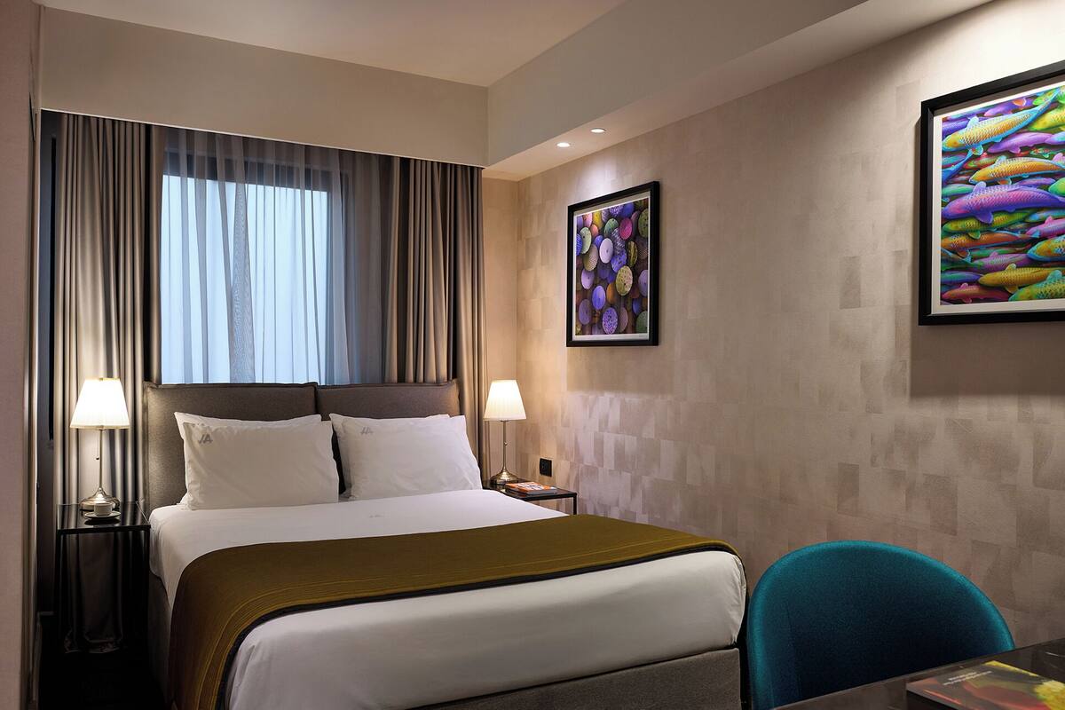 A hotel room with a bed, two bedside tables with lamps, a chair, and two framed artworks on the wall.
