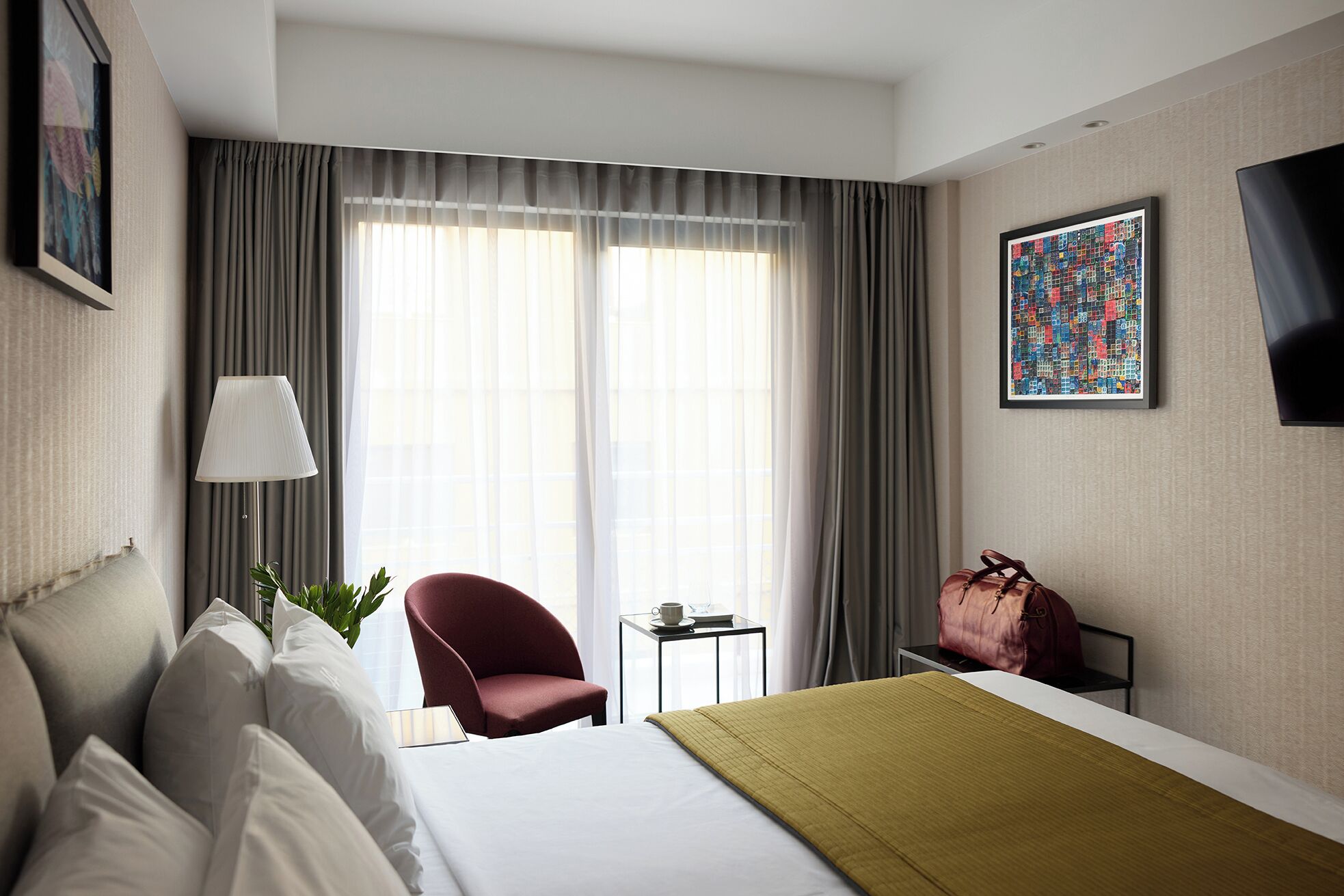 A hotel room with a bed, a chair, a small table with a lamp, a TV, and a painting on the wall.