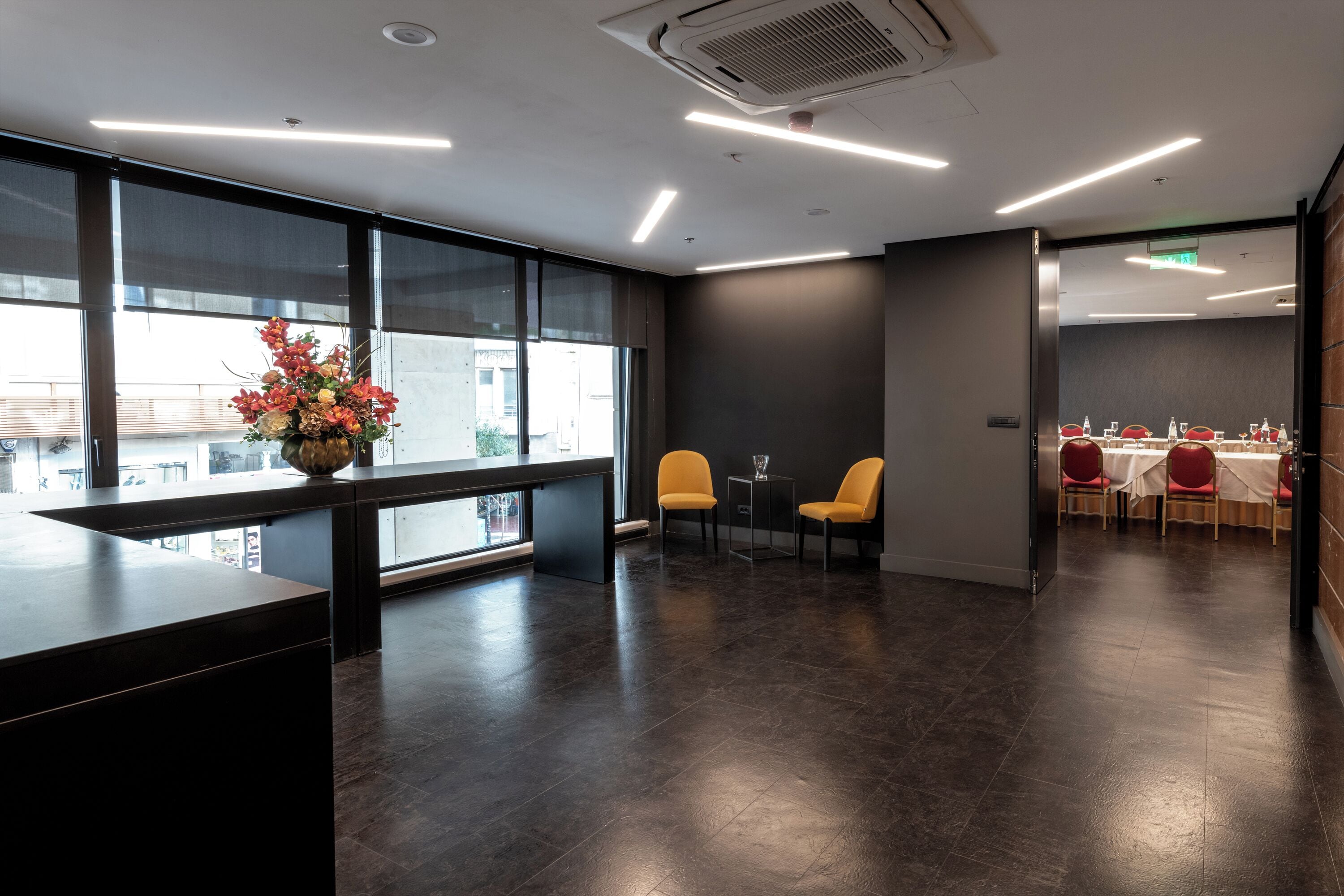 A modern hotel lobby with a reception desk, large windows, and a dining area with red chairs and a table.