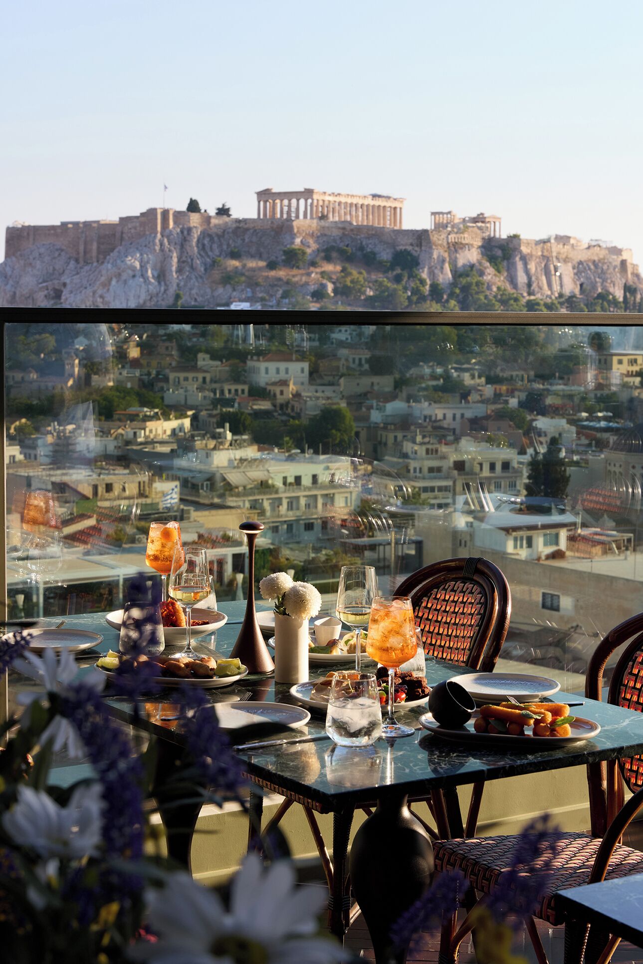 A table set with glasses, a vase with flowers, and plates of food, overlooking a cityscape with ancient ruins.