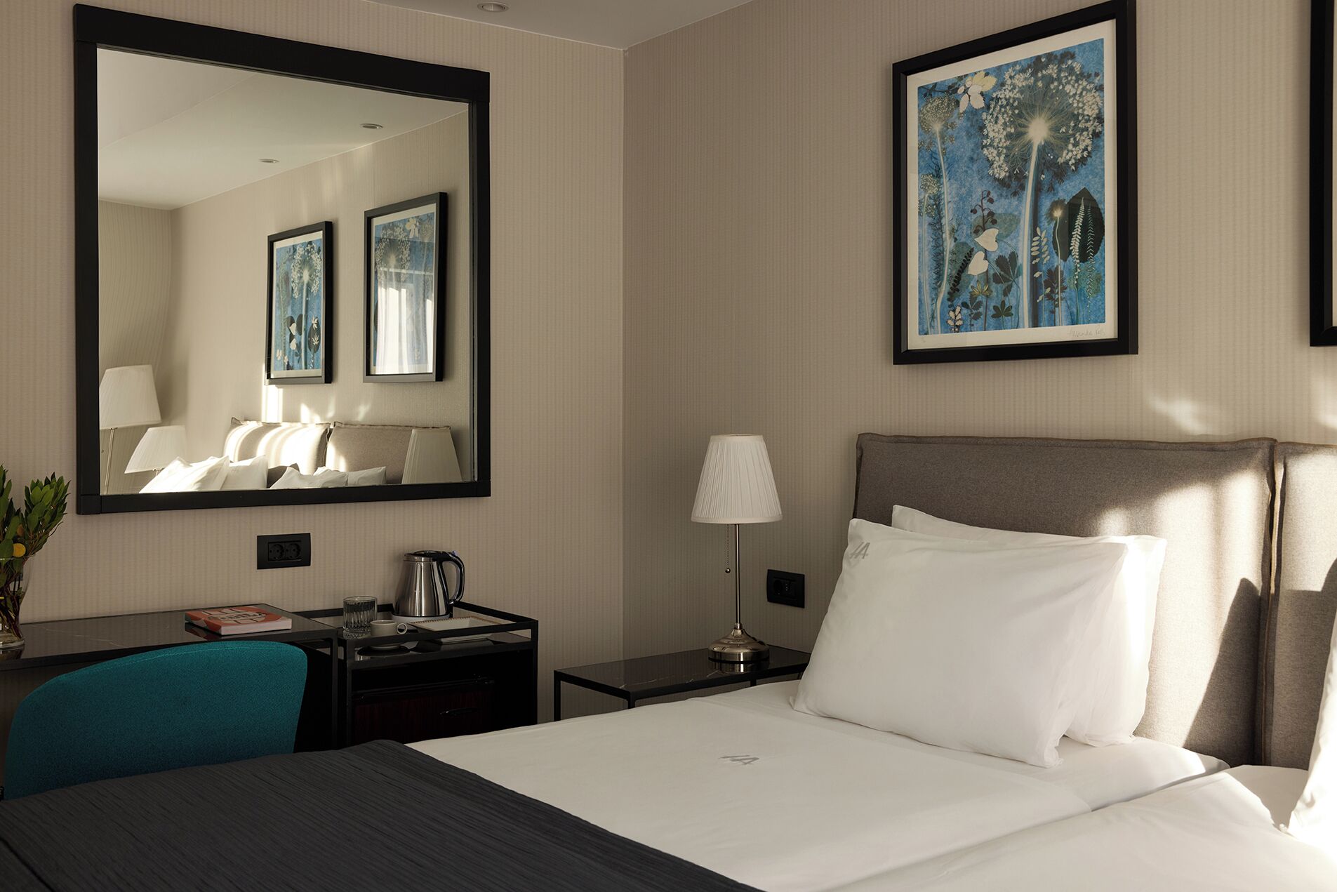 Une chambre d’hôtel avec un grand lit, un bureau avec une lampe, une machine à café et un tableau botanique encadré accroché au mur.