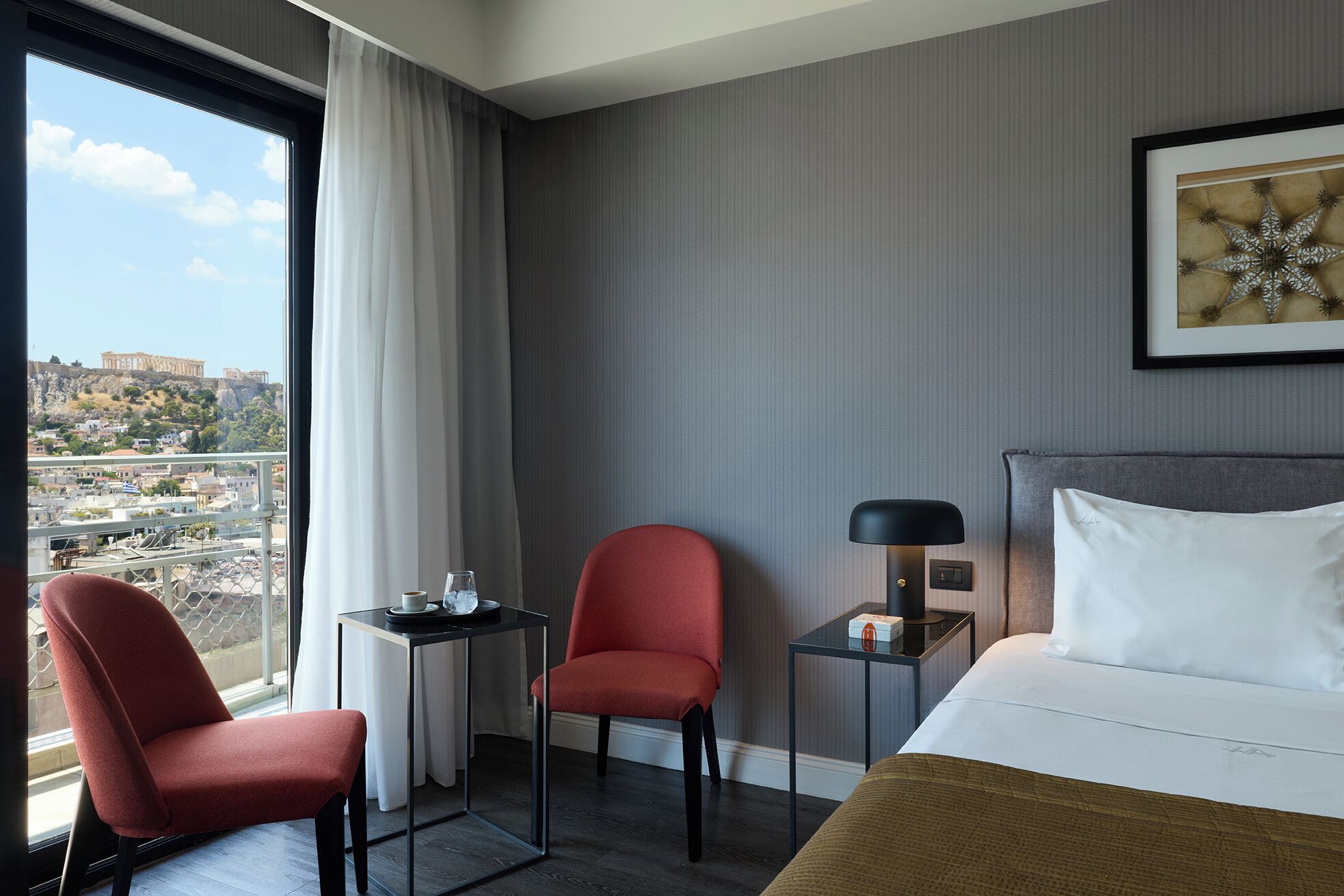 Ein Hotelzimmer mit einem Bett, einem roten Sessel, einem kleinen Tisch mit Lampe und Blick auf die Stadt.