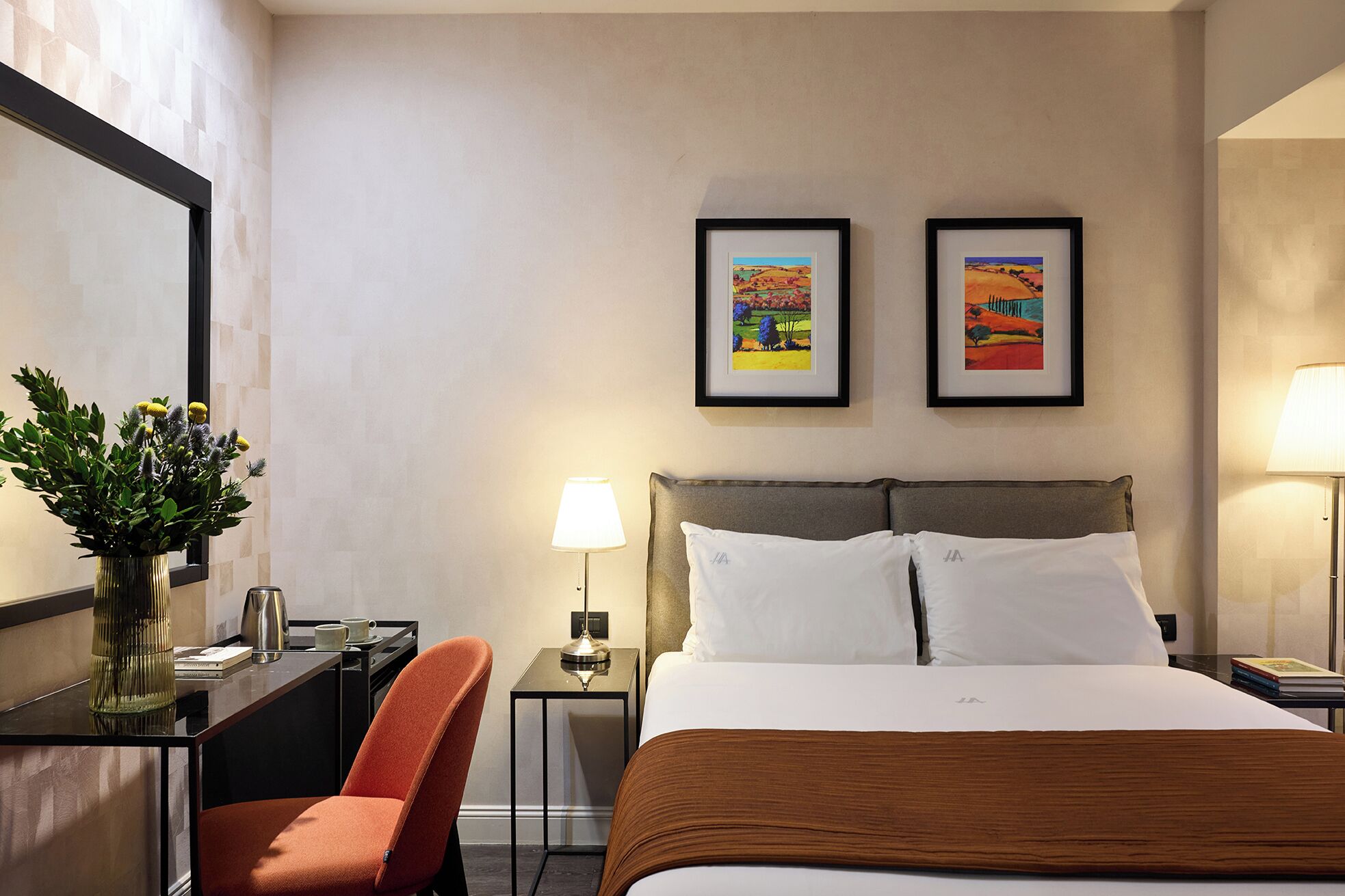 A hotel room with a bed, two framed artworks on the wall, a desk with a chair, a bedside table with a lamp, and a decorative plant.