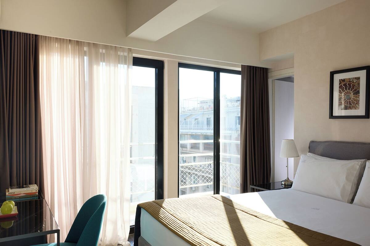 A hotel room with a bed, a desk with a lamp, a chair, and a view of buildings through large windows.