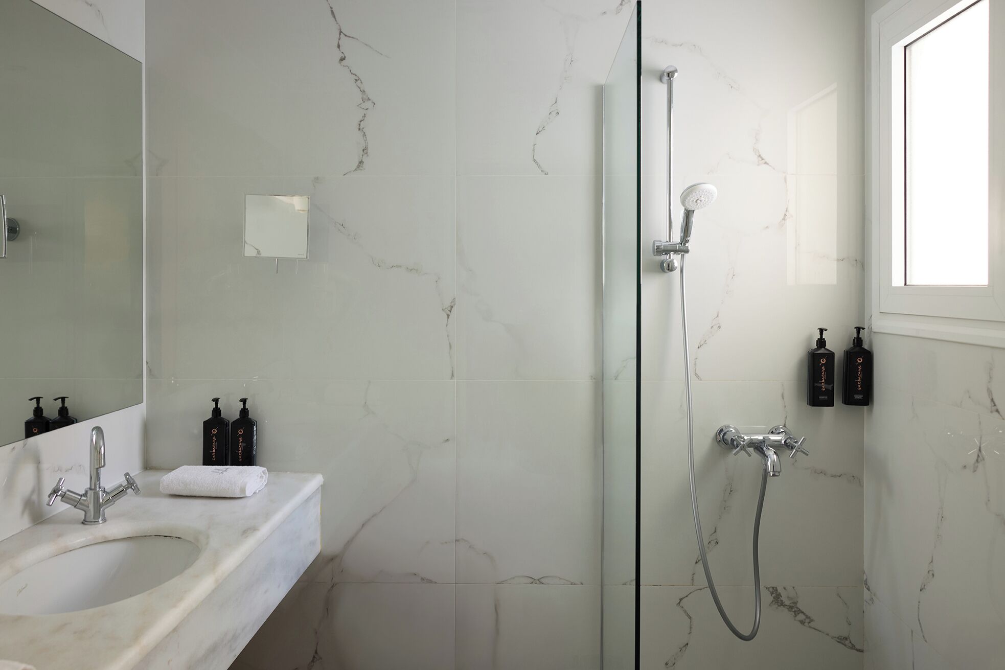 A bathroom with a marble wall, a glass shower enclosure, a sink with a mirror, and toiletries on the counter.