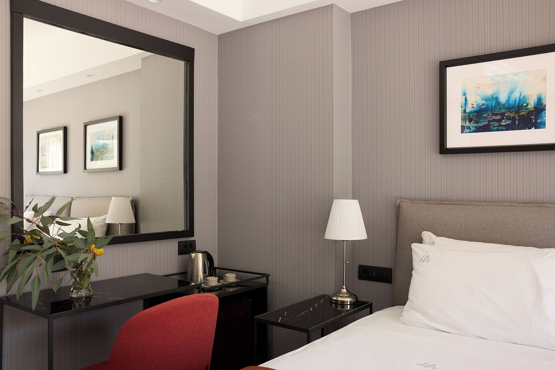 A hotel room with a bed, a desk, a chair, a large mirror, a lamp, and a painting on the wall.
