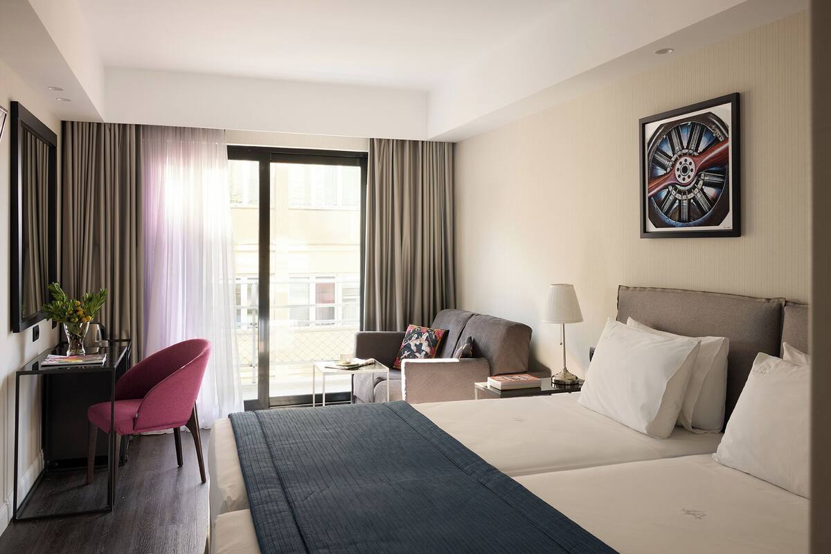A hotel room with a bed, a desk with a chair, a sofa, and a large window with curtains.