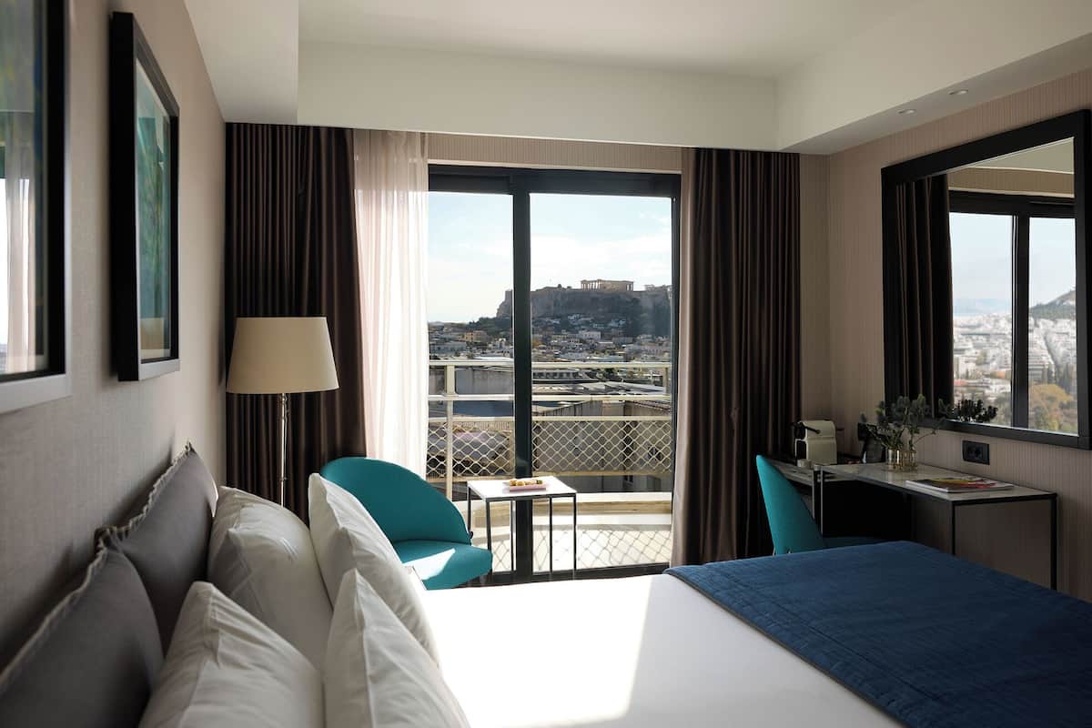 A hotel room with a bed, a desk, a chair, and a balcony with a city view.