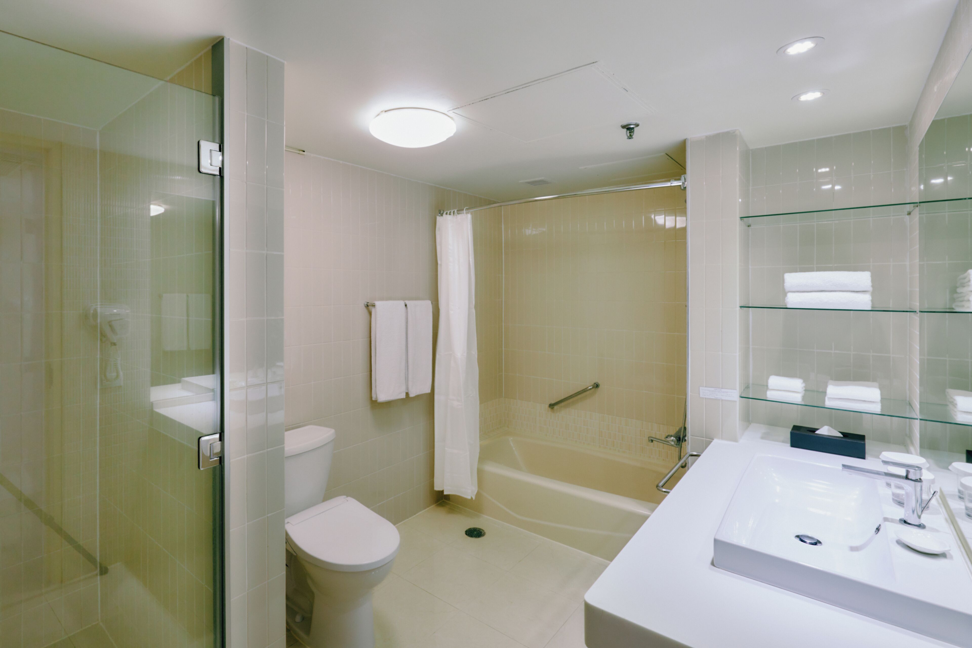 Un baño moderno con ducha envuelta en vidrio, inodoro blanco, bañera y tocador con espejo y cubierta de vidrio.