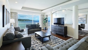A modern living room with a sofa, coffee table, and TV, large windows with a view of the sea, and a balcony.