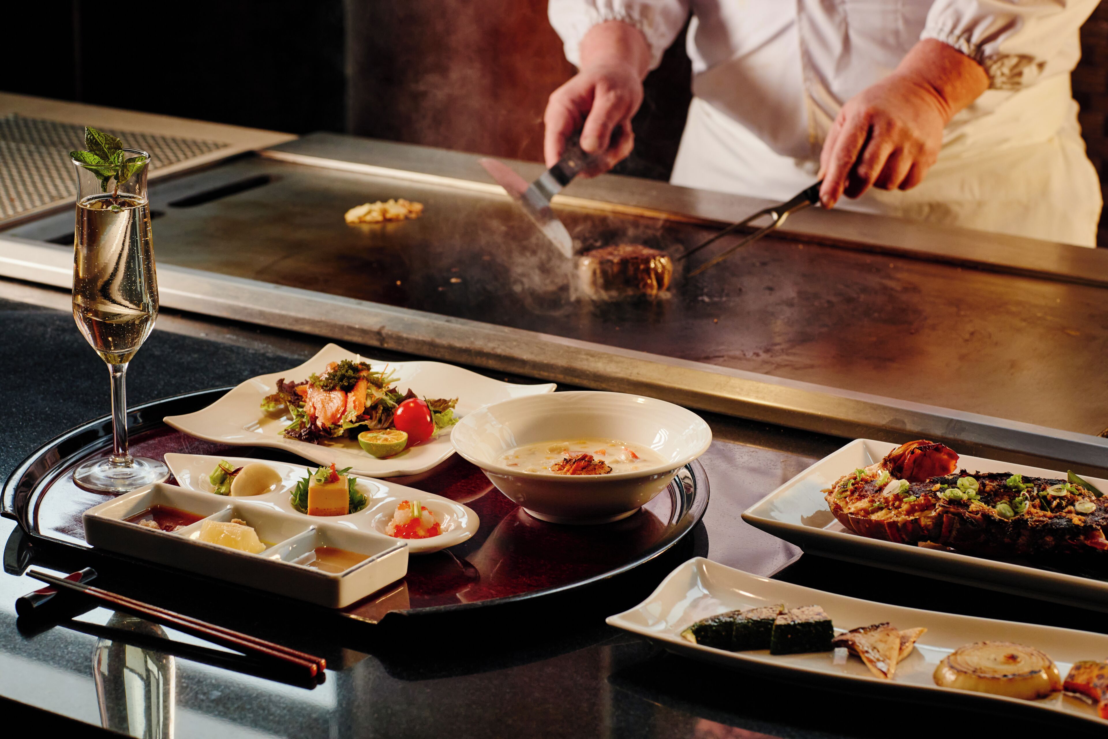 A dining table with various dishes, a glass of champagne, and a person cooking.