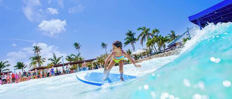 A person surfing on a wave at a water park with palm trees and a clear sky in the background.