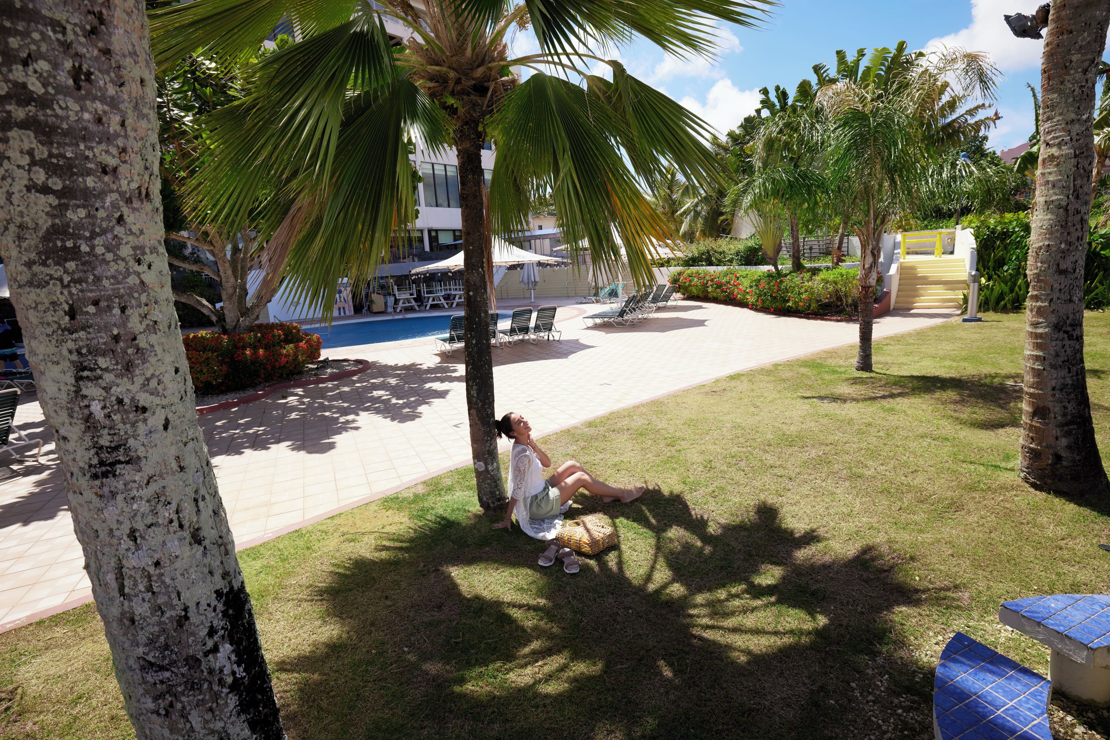 Un espace piscine avec des chaises longues, des palmiers et une personne assise sur l’herbe.