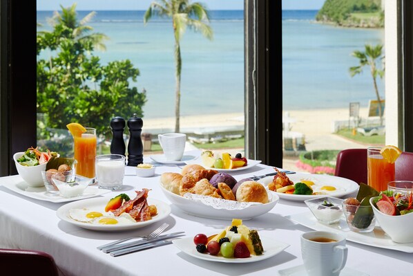 A breakfast table with various dishes, a view of the beach, and a tropical landscape.