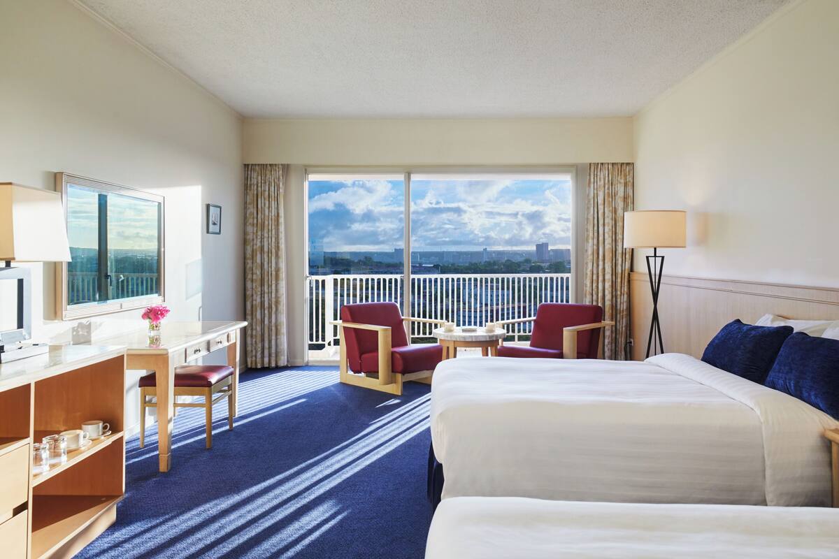 A hotel room with a large bed, a desk with a chair, a small table, and a view of the city through the balcony doors.