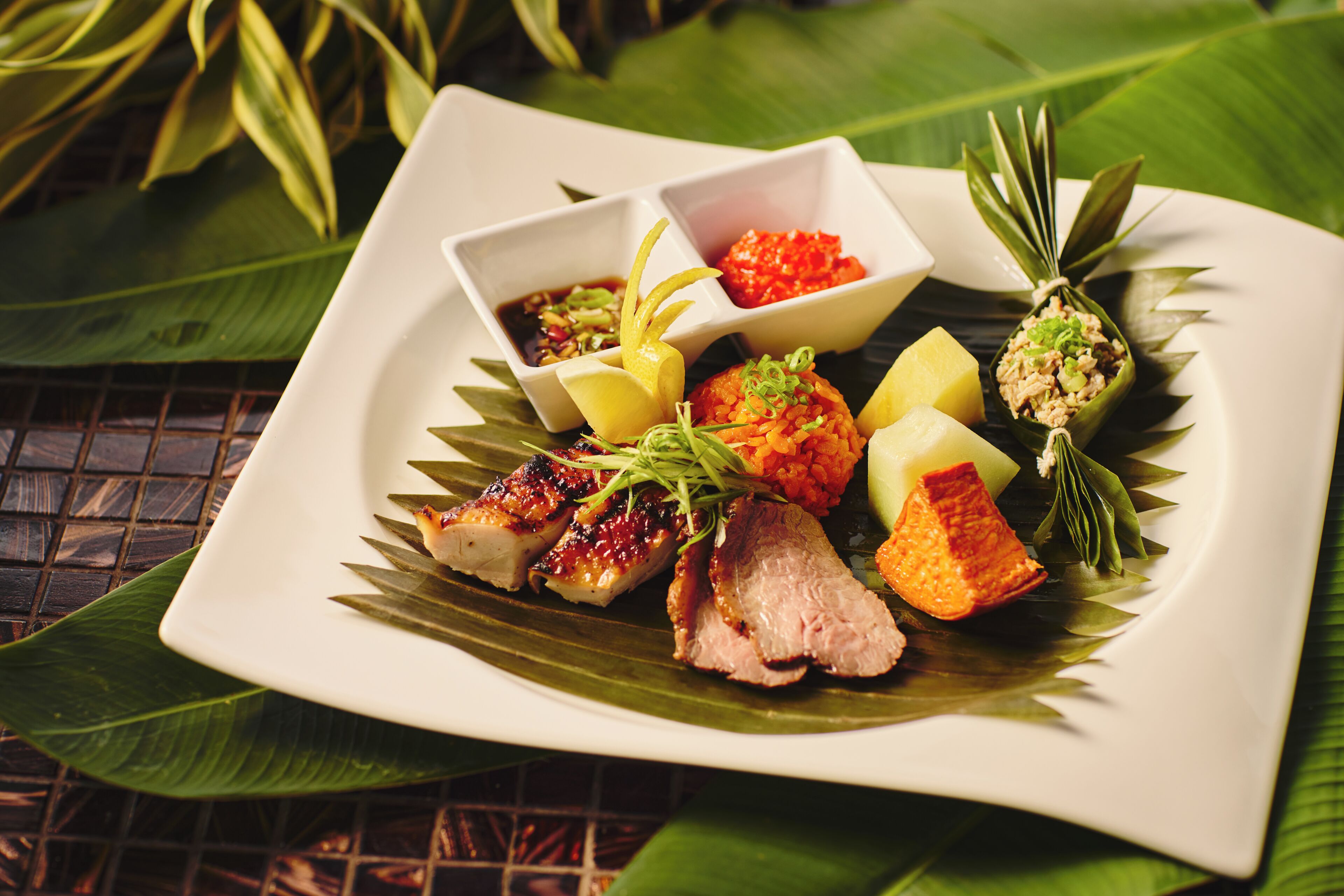 A square white plate with grilled meat, pineapple, and two small bowls of sauce, set on a table with green leaves.