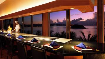 A restaurant with a long counter, multiple chairs, and a view of a body of water at dusk.