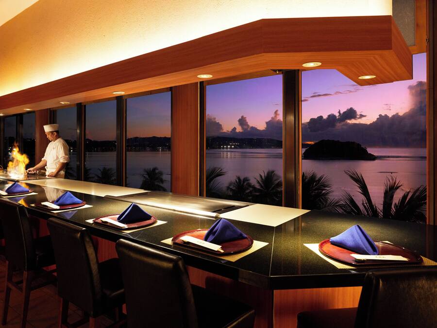 A restaurant with a long counter, multiple chairs, and a view of a body of water at dusk.