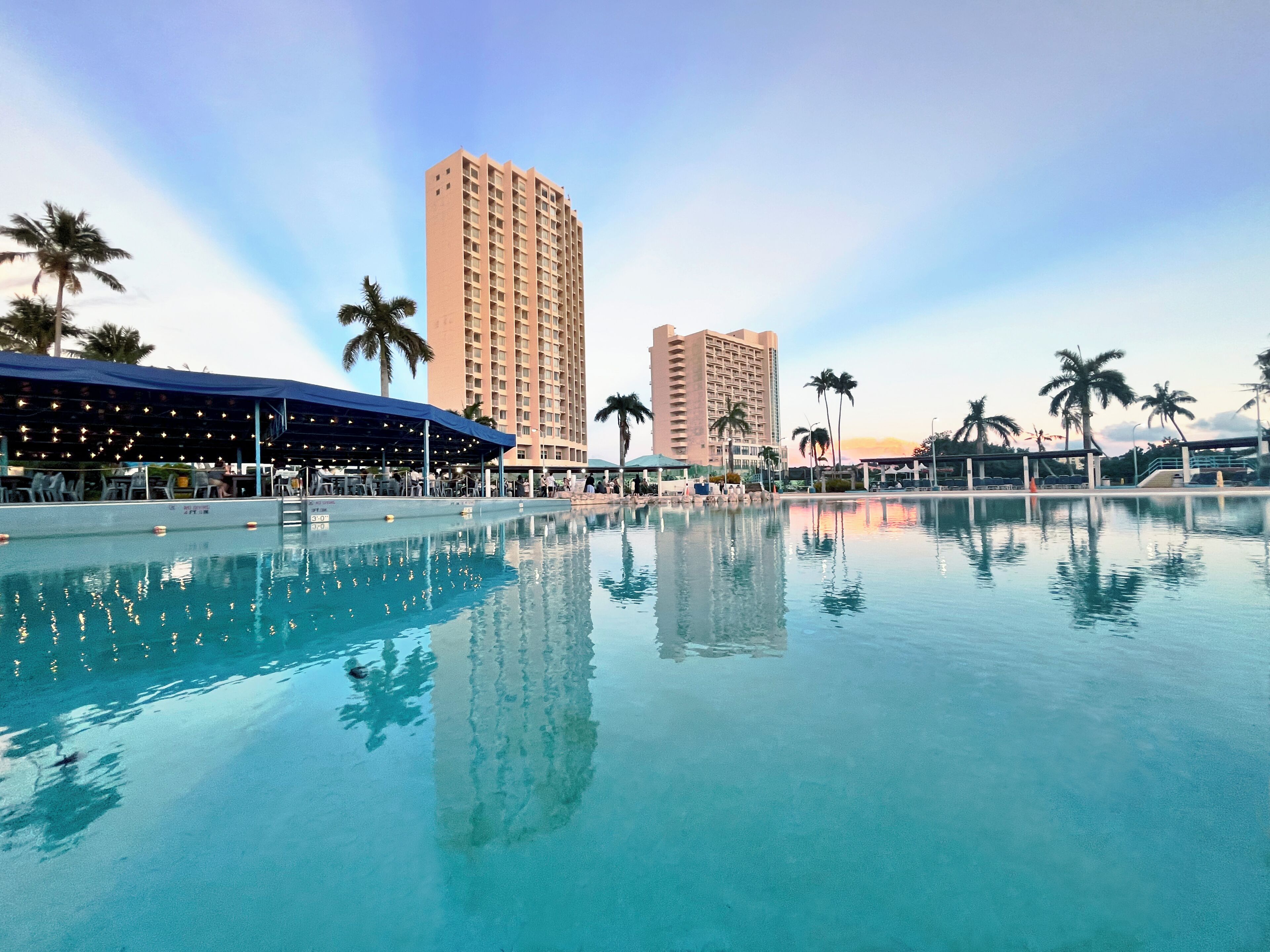 A swimming pool with clear water, surrounded by palm trees and a tall hotel building.