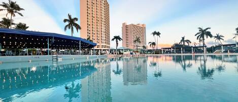 A swimming pool with clear water, surrounded by palm trees and a tall hotel building.