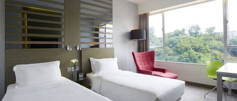 A hotel room with two beds, a red chair, a green chair, a desk, and a large window with a view of trees.