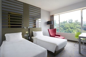Habitación de hotel con dos camas, una silla roja, una silla verde, un escritorio y un ventanal con vistas a árboles.