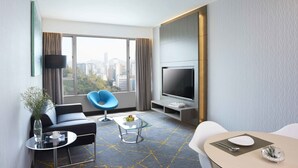 A modern hotel room with a large window, a flat-screen TV, a sofa, a blue chair, a dining table with a white table runner, and a vase with flowers. - The Cityview - Chinese YMCA of Hong Kong (Kowloon)