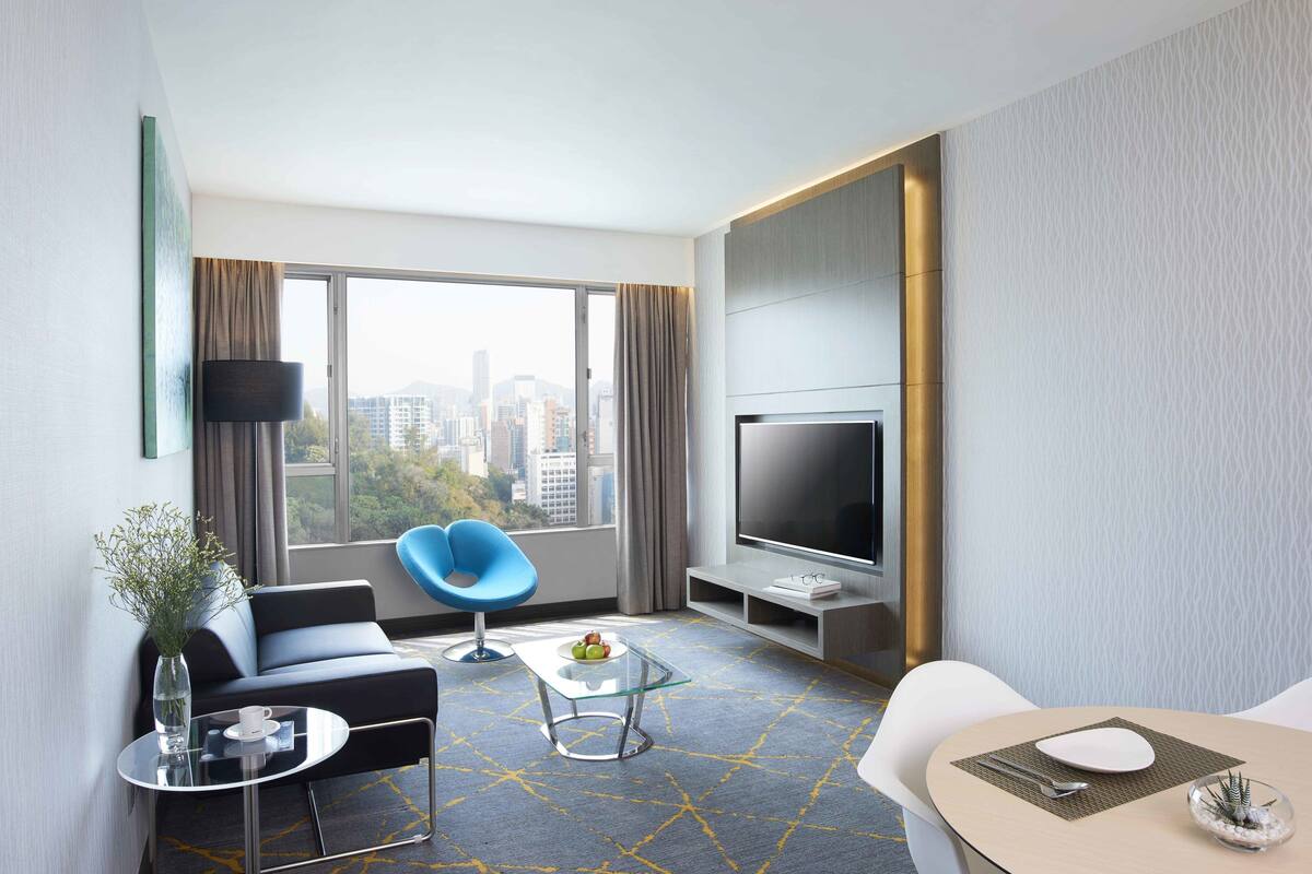 A modern hotel room with a large window, a flat-screen TV, a sofa, a blue chair, a dining table with a white table runner, and a vase with flowers.