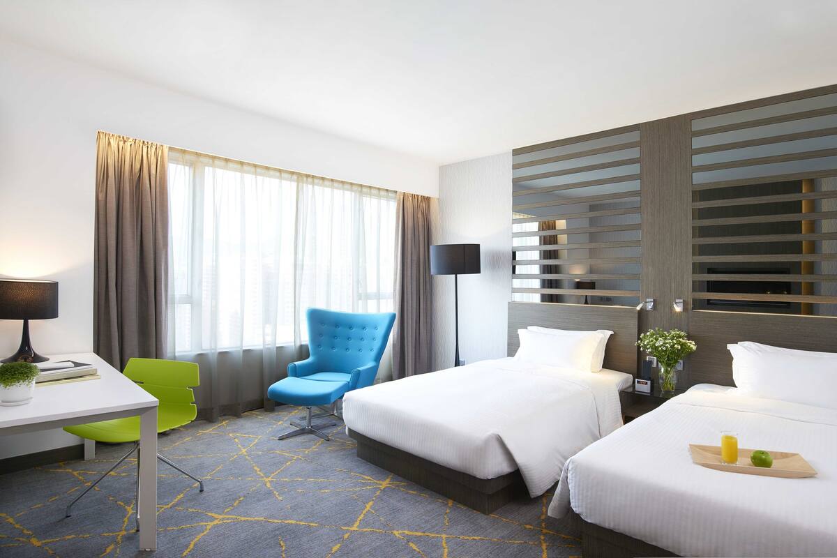A hotel room with two beds, a desk with a chair, a lamp, and a window with curtains.