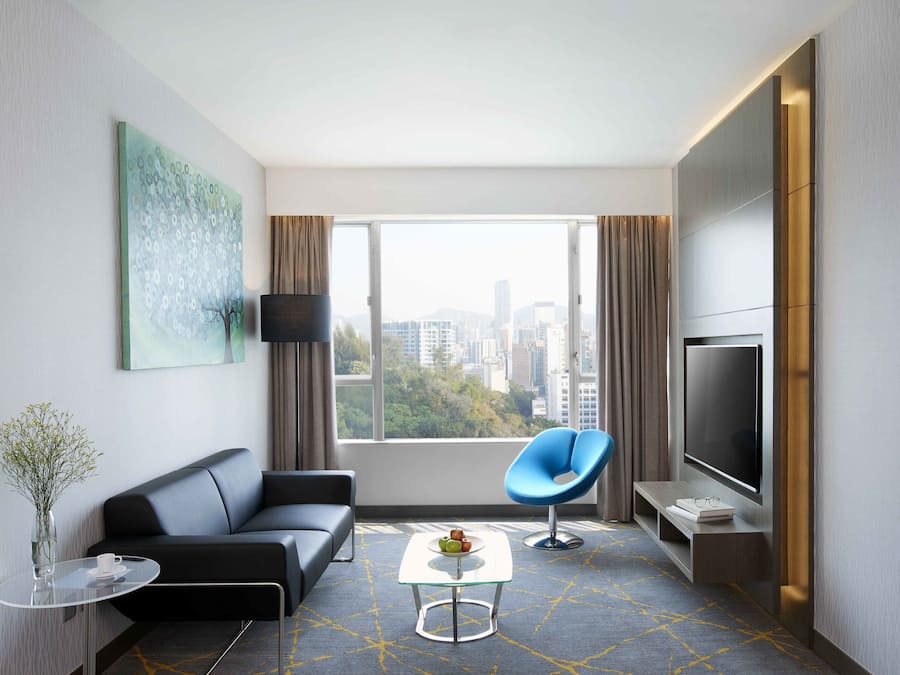 A modern hotel room with a sofa, a blue chair, a coffee table, a TV, and a large window offering a city view.
