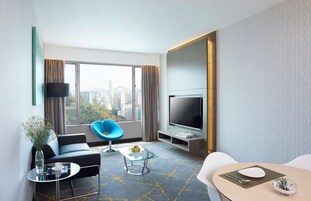 A modern hotel room with a city view, a flat-screen TV, a sofa, a coffee table, and a dining area.