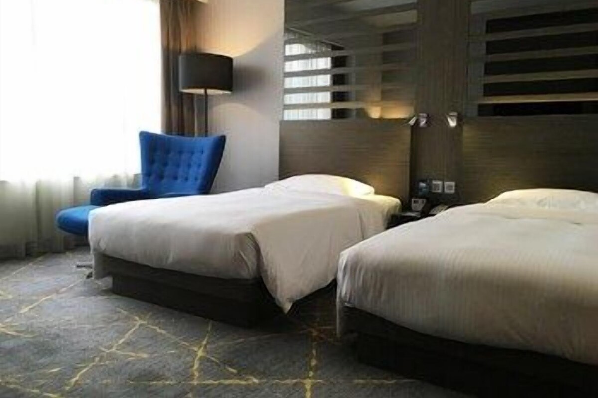 Una habitación de hotel con dos camas, una silla azul, una lámpara y una alfombra estampada.