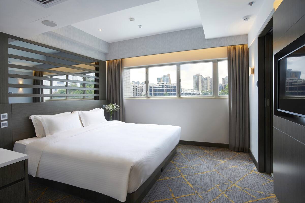 A modern hotel room with a large bed, a TV, a city view, and a window with curtains.