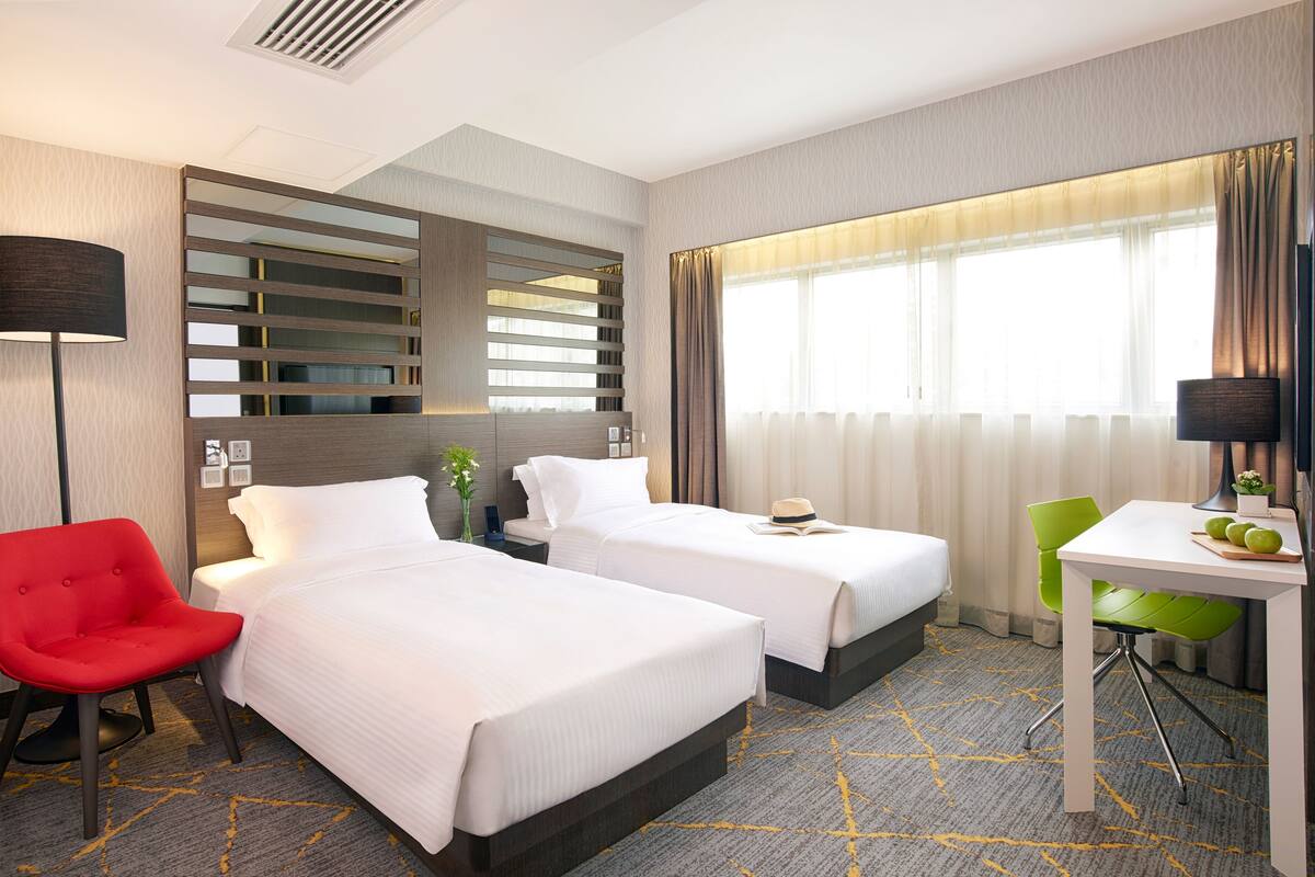 A hotel room with two beds, a desk, a chair, and a lamp.