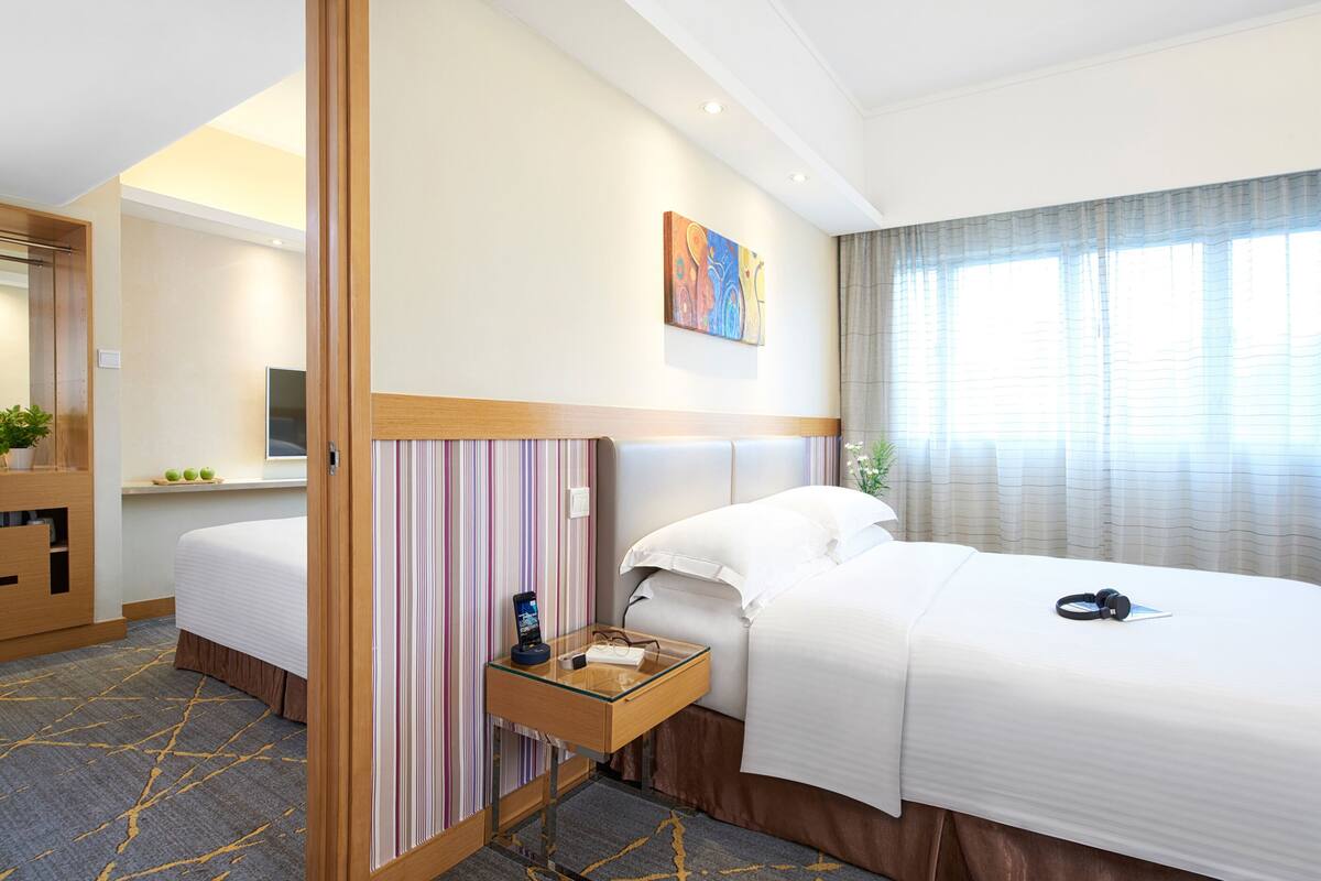 A hotel room with two beds, a bedside table with a phone and glasses, a painting on the wall, and a window with curtains.