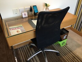 Un espacio de trabajo en un hotel con un escritorio de vidrio, una silla de oficina de malla negra, un teléfono móvil cargando, un cartel de prohibición de fumar, una revista CityLife y un contenedor de reciclaje.