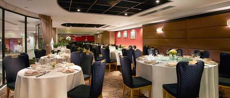 A hotel dining area with round tables, dark upholstered chairs, and a patterned carpet.
