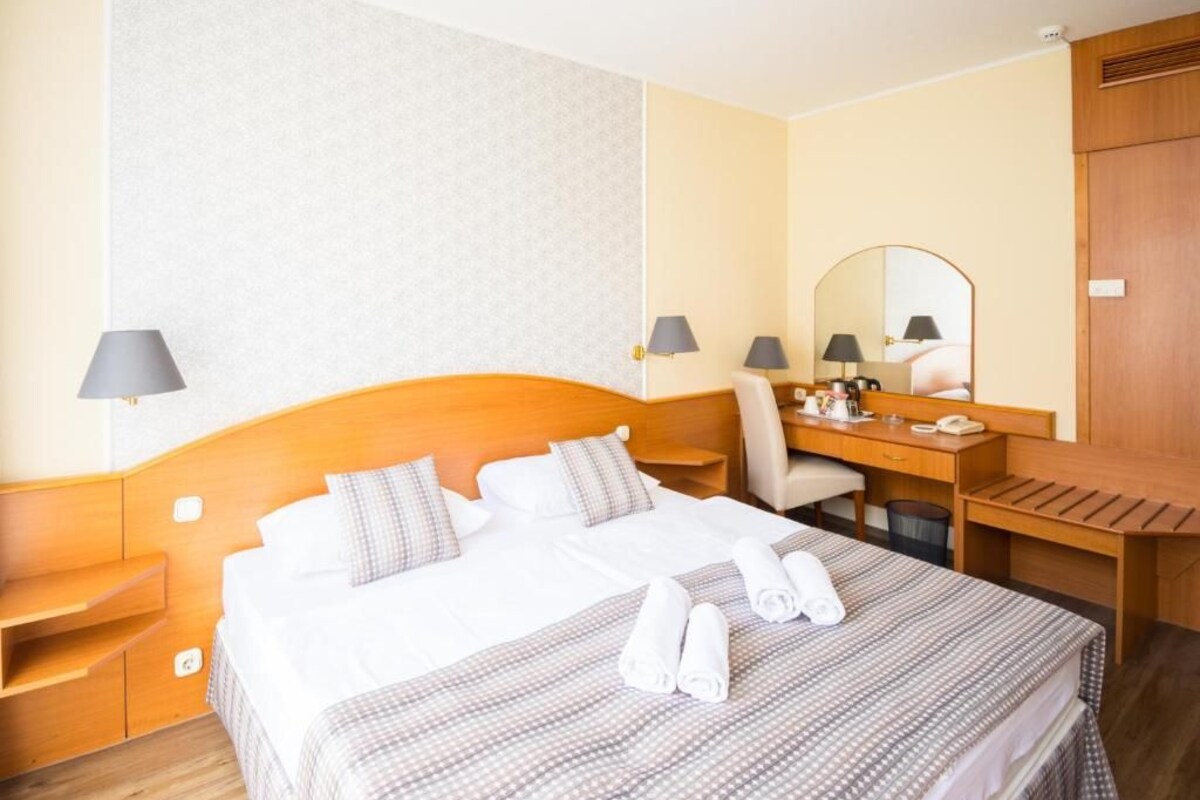 A hotel room with a bed, a desk, a chair, a mirror, and a lamp.