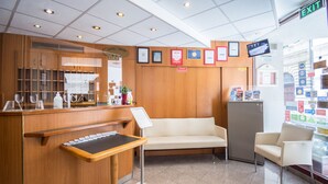 A hotel reception area with a wooden counter, a waiting area with a sofa, a small refrigerator, and a television mounted on the wall. - Hotel Orion (Budapest)