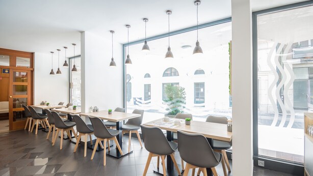 A modern dining area with long tables and chairs, large windows with a view of buildings, pendant lighting, and a tiled floor. - Hotel Orion (Budapest)