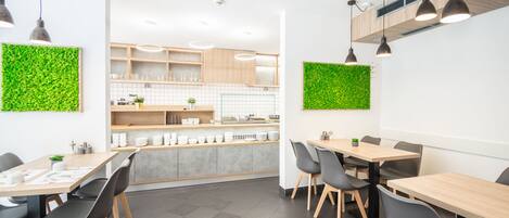 A modern dining area with wooden tables and black chairs, a kitchen in the background, and green wall decorations.