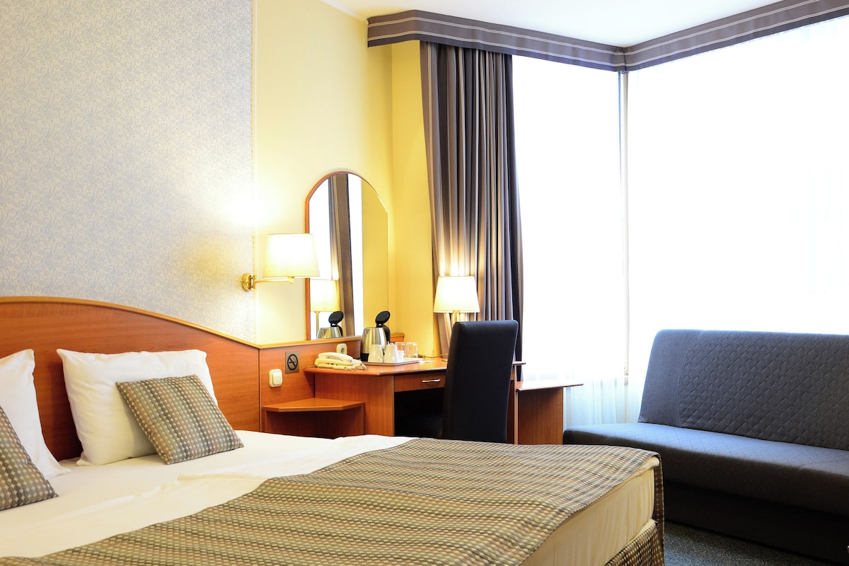 A hotel room with a large bed, a desk with a chair, a sofa, and a window with curtains.