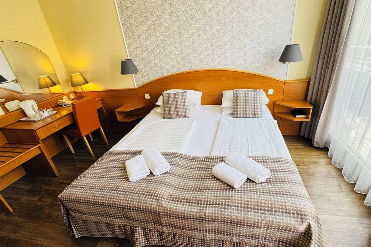 Habitación de hotel con una cama grande, dos mesitas de noche, un escritorio con una silla, un espejo y una ventana con cortinas.