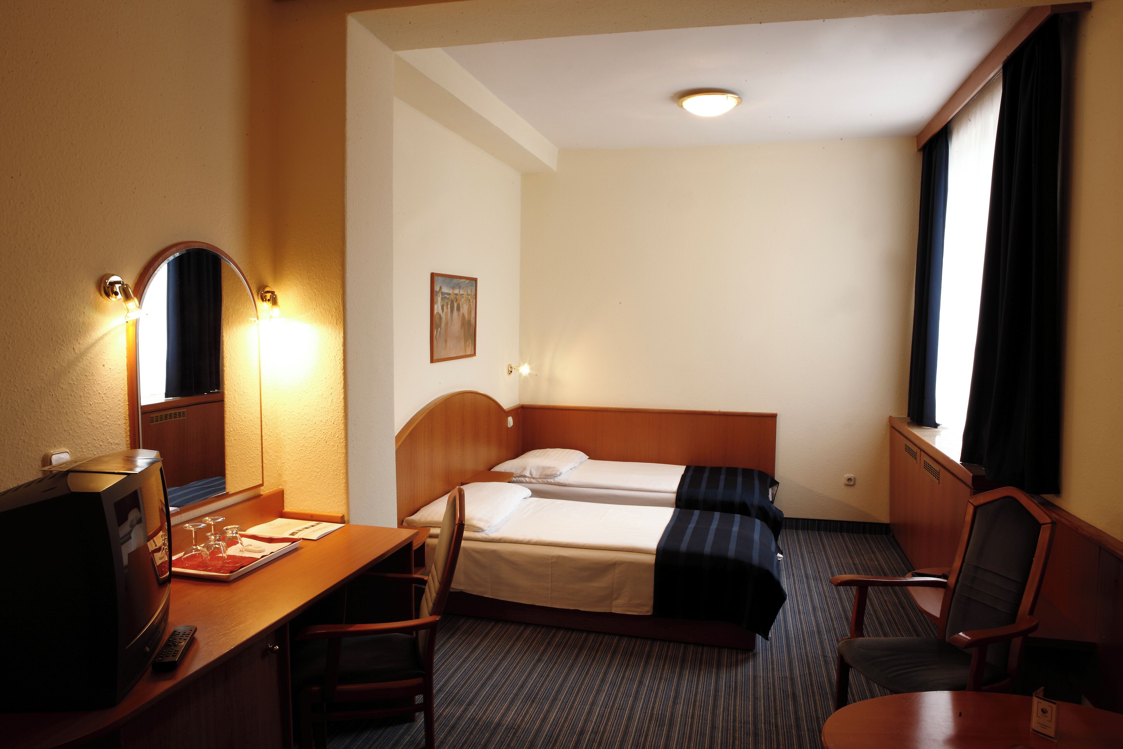 A hotel room with a bed, a desk with a computer and a TV, a chair, and a window with curtains.