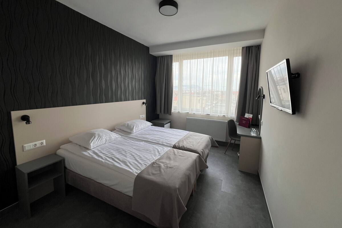 Une chambre d’hôtel avec deux lits, un bureau, une chaise, une fenêtre avec des rideaux et un téléviseur fixé au mur.