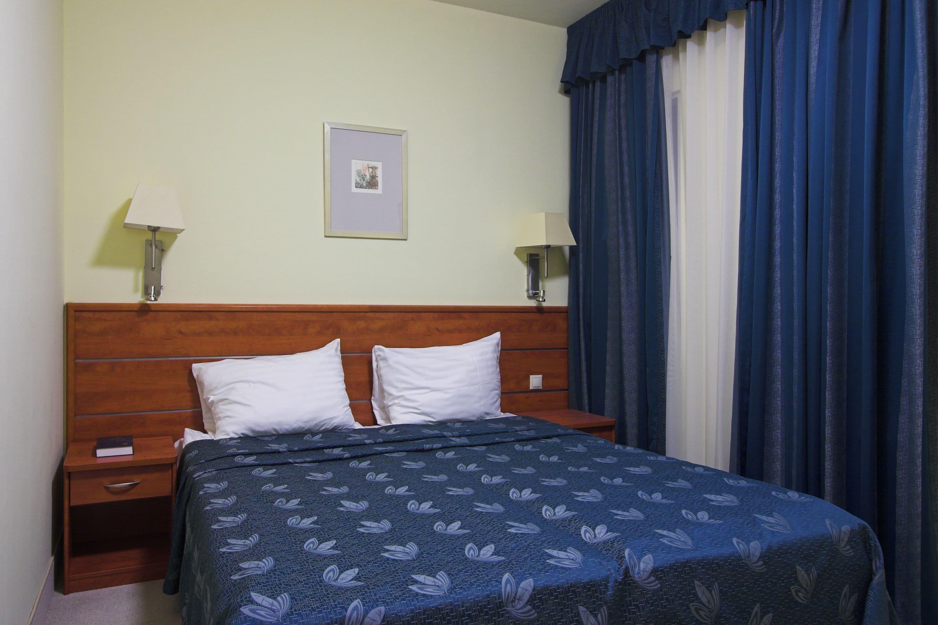 A hotel room with a wooden headboard, two bedside tables with lamps, a bed with a patterned bedspread, and a framed picture on the wall.