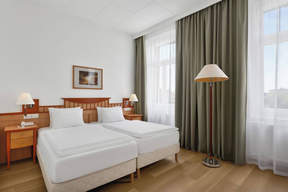 Habitación de hotel con dos camas, cabecero de madera, mesita de noche con lámpara, un cuadro enmarcado en la pared y un ventanal con cortinas.