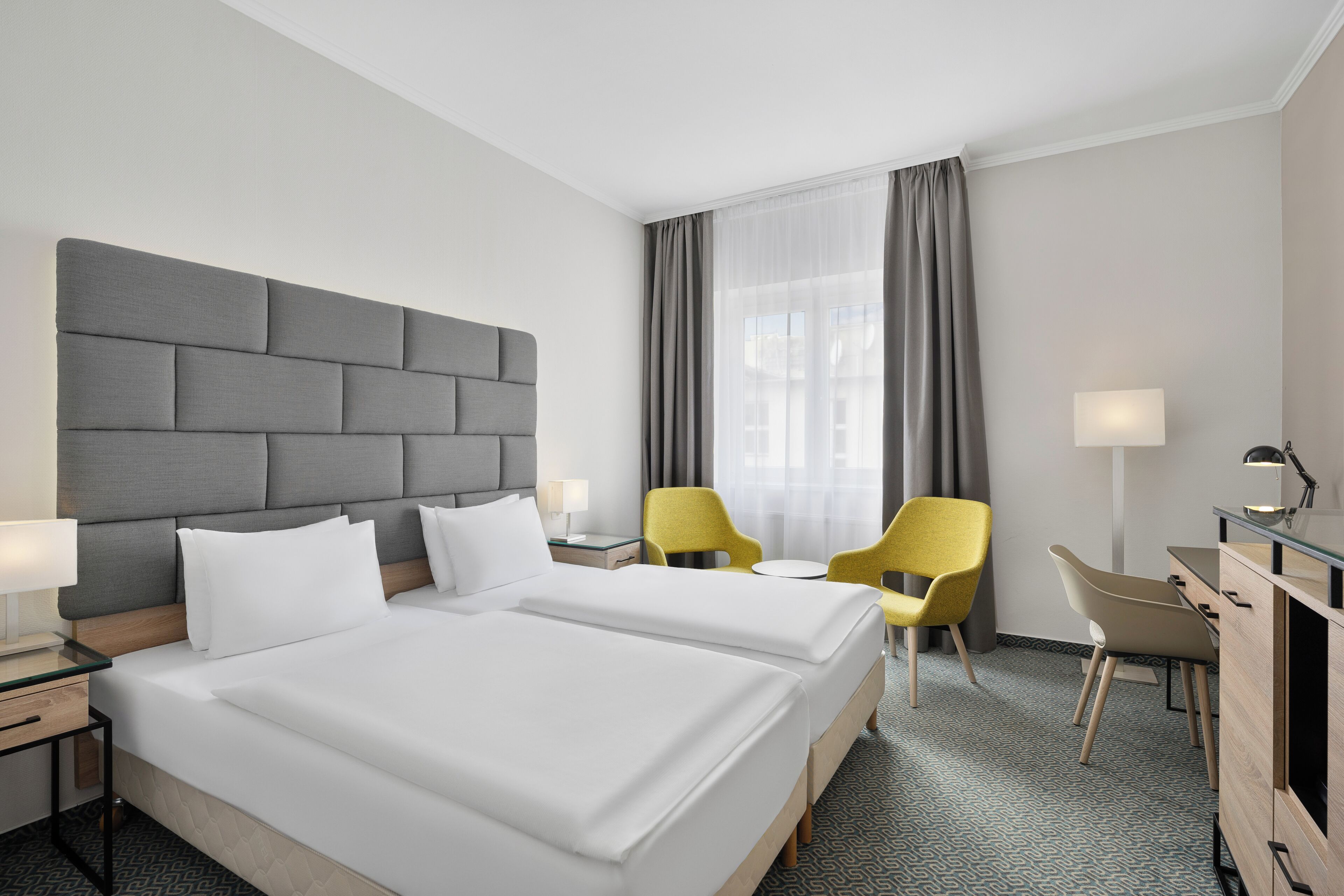 A hotel room with two beds, a desk, a chair, a lamp, and a window with curtains.
